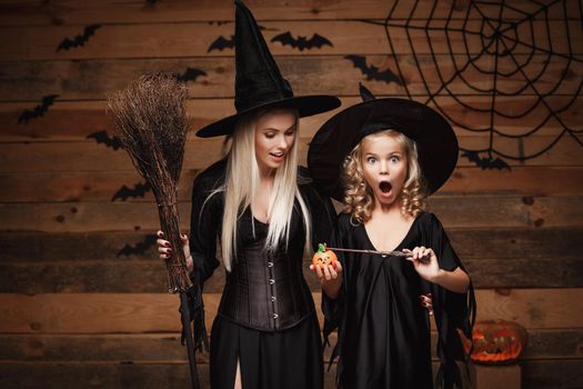 Halloween Concept: beautiful caucasian mother and her daughter in witch costumes enjoy using magic with magic wand to halloween pumpkin jar over bats and spider web on Wooden studio background.