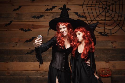 Halloween Concept - Beautiful caucasian mother and her daughter with long red hair in witch costumes taking a selfie with smartphone ready for celebrating Halloween