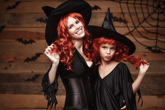 Halloween Concept: Beautiful caucasian mother and her daughter with long red hair in witch costumes celebrating Halloween posing with over bats and spider web on Wooden studio background