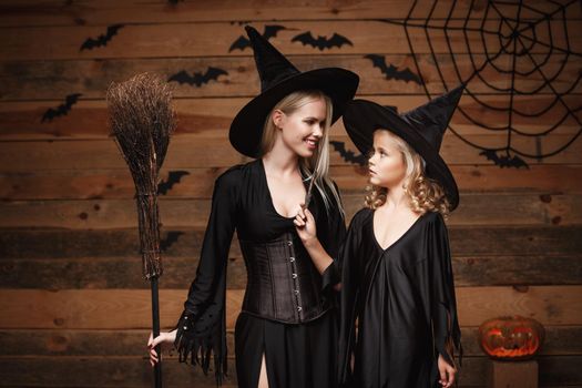 Halloween Concept - cheerful mother and her daughter in witch costumes celebrating Halloween posing with curved pumpkins over bats and spider web on Wooden studio background