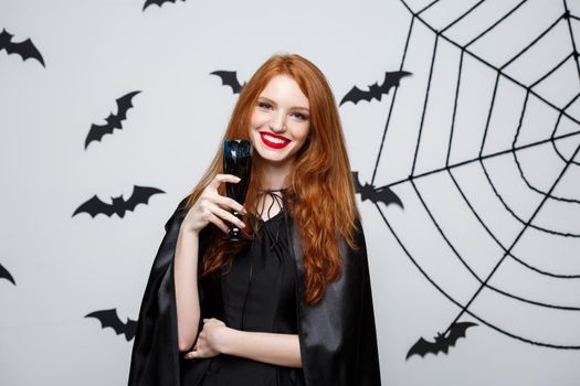 Halloween witch concept - Happy Halloween Witch holding glass of bloody red wine over dark grey studio background with bat and spider web