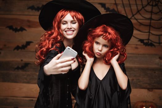 Halloween Concept - Beautiful caucasian mother and her daughter with long red hair in witch costumes taking a selfie with smartphone ready for celebrating Halloween