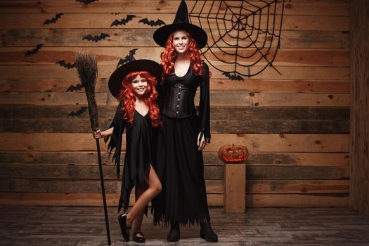 Halloween Concept: Beautiful caucasian mother and her daughter with long red hair in witch costumes celebrating Halloween posing with over bats and spider web on Wooden studio background