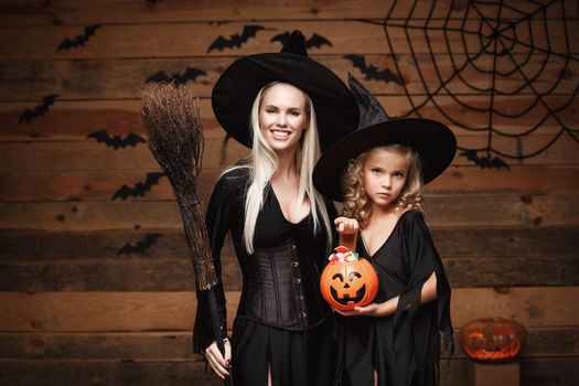 Halloween Concept: beautiful caucasian mother and her daughter in witch costumes celebrating Halloween with sharing Halloween candy and sweet over bats and spider web on Wooden studio background.