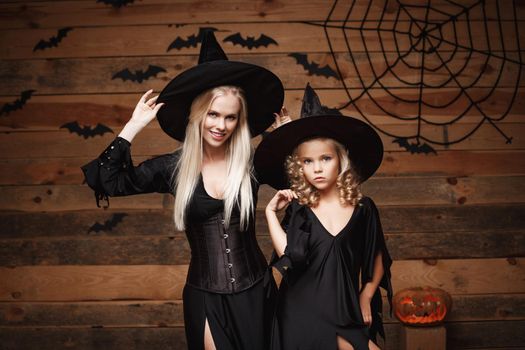 Halloween Concept - cheerful mother and her daughter in witch costumes celebrating Halloween posing with curved pumpkins over bats and spider web on Wooden studio background