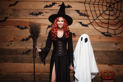 Halloween Concept: Witch mother and little white ghost doing trick or treat celebrating Halloween posing with curved pumpkins over bats and spider web on Wooden studio background
