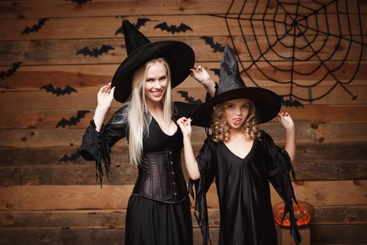 Halloween Concept - cheerful mother and her daughter in witch costumes celebrating Halloween posing with curved pumpkins over bats and spider web on Wooden studio background