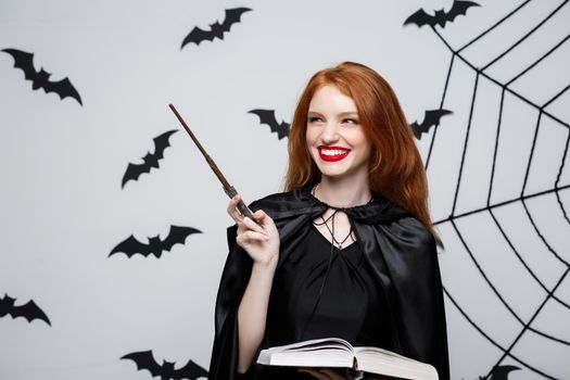 Halloween Concept - Beautiful Witch playing with magic stick and magic book on grey background