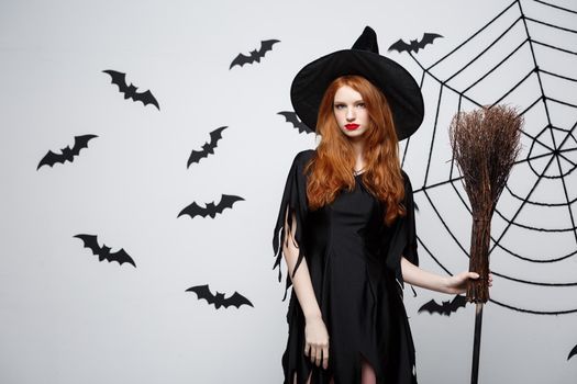 Halloween Witch Concept - Portrait of beautiful young witch with broomstick over grey wall with bat and spider web background