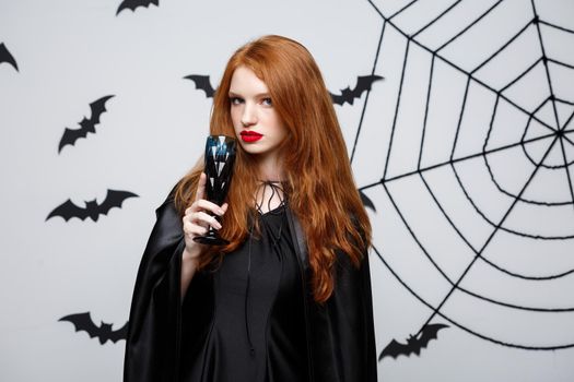 Halloween witch concept - Happy Halloween Witch holding glass of bloody red wine over dark grey studio background with bat and spider web