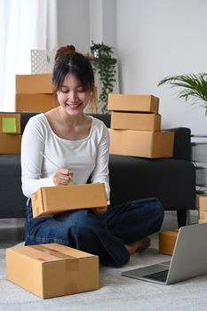 Smiling young asian woman sitting on floor preparing parcel boxes of product for delivery and writing address on cardboard box.