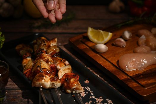 Man hand sprinkling salt and seasoning on grilled skewers of chicken and vegetables.
