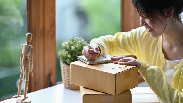 Focused young woman online seller writing address on cardboard box while preparing parcel boxes of product for delivery.