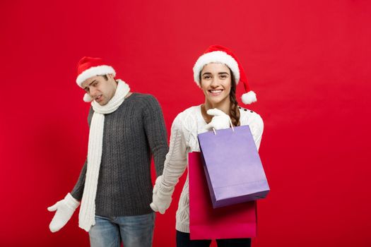 Christmas Concept - Beautiful girlfriend force her boyfriend to go shopping over red christmas background.