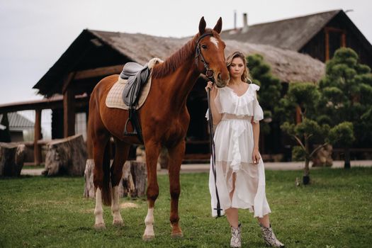 Beautiful girl in a white sundress next to a horse on an old ranch.