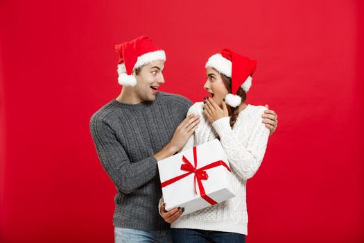 Christmas Concept - Young woman covering man's eyes with hand and giving surprise big gift. Isolated on Red background.