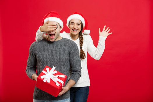 Christmas Concept - Young woman covering man's eyes with hand and giving surprise big gift. Isolated on Red background.