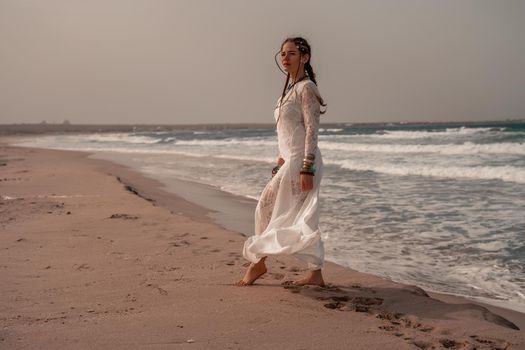 Model in boho style in a white long dress and silver jewelry on the beach. Her hair is braided, and there are many bracelets on her arms