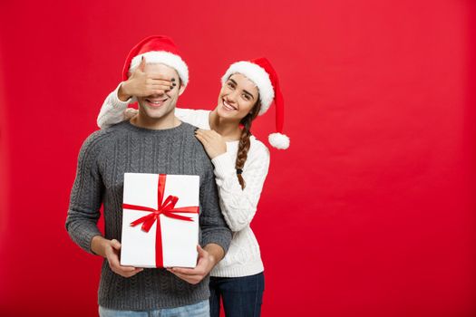 Christmas Concept - Young woman covering man's eyes with hand and giving surprise big gift. Isolated on Red background.