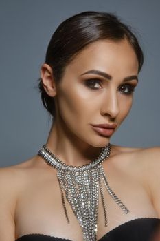 Close up portrait of a beautiful brunette model female with glowing perfect skin, professional evening make-up and plump lips, wearing a black bra and necklace. She is looking at the camera while posing against a gray studio background. Copy space, soft effect, vintage.