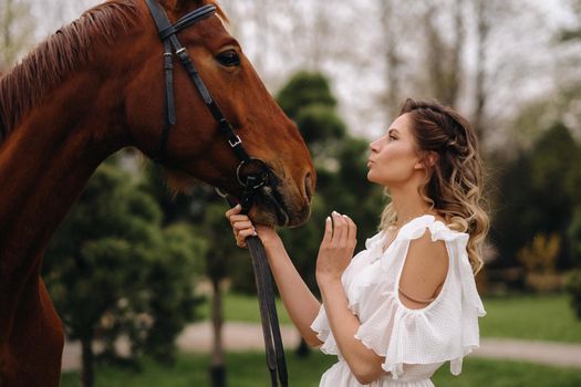 Beautiful girl in a white sundress next to a horse on an old ranch.