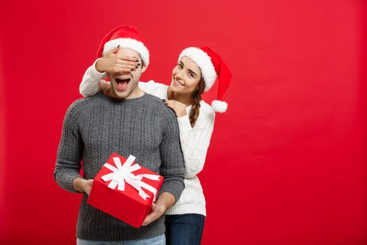 Christmas Concept - Young woman covering man's eyes with hand and giving surprise big gift. Isolated on Red background.