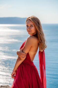 Smiling young woman in a red dress looks at the camera. A beautiful tanned girl enjoys her summer holidays at the sea. Portrait of a stylish carefree woman laughing at the ocean