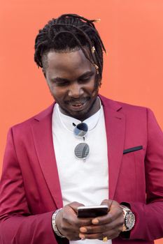 vertical portrait of a stylish african businessman in suit typing a message on his smart phone in front of a red background, copy space for text, concept of technology and communication