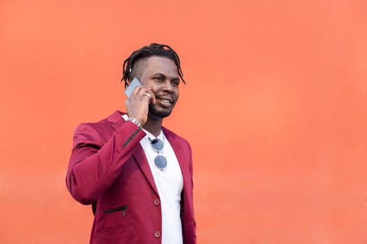 stylish black businessman in suit smiling and talking on phone in front of red background, copy space for text, concept of technology and communication