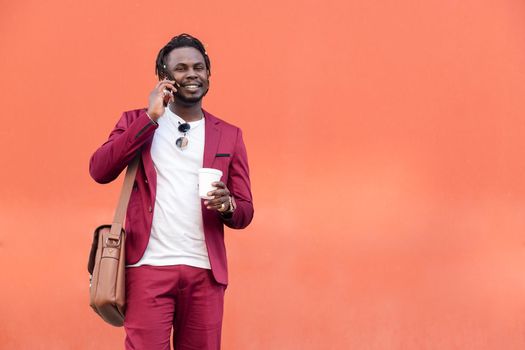 stylish black businessman dressed in suit with briefcase and coffee talks on smart phone in front of red background, copy space for text, concept of technology and communication