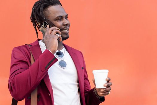 portrait of a stylish african businessman dressed in suit smile with a coffee on his hand while talks by phone in front of red background, copy space for text, concept of technology and communication