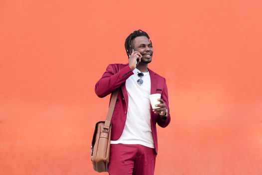 stylish african businessman dressed in suit smiles with a coffee on his hand while talks by phone in front of red background, copy space for text, concept of technology and communication