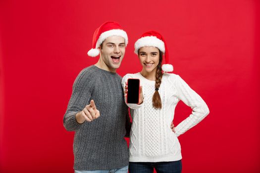 Christmas concept - Happy young couple in christmas sweaters pointing finger with mobile phone.