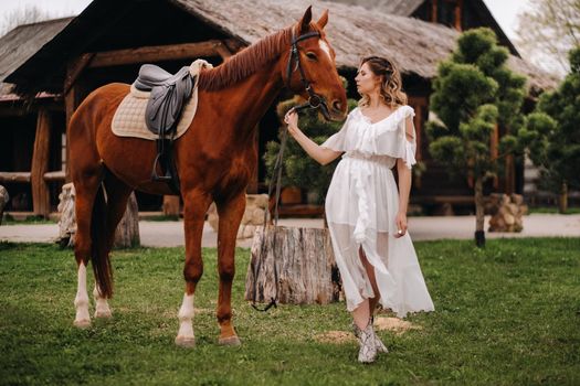 Beautiful girl in a white sundress next to a horse on an old ranch.