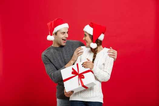 Christmas Concept - Young woman covering man's eyes with hand and giving surprise big gift. Isolated on Red background.