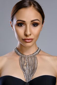 Close up portrait of a beautiful brunette fashion model woman with glowing perfect skin, professional evening make-up and plump lips, wearing a black bra and necklace. She is looking at the camera while posing against a gray studio background. Copy space.