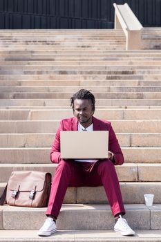 elegant african businessman works with his laptop sitting on a staircase in the city, technology and remote work concept, copy space for text