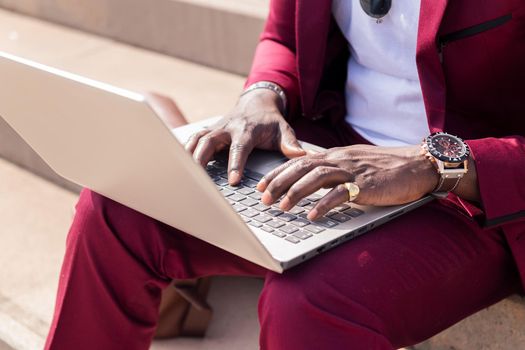 detail of the hands of an unrecognizable black man working with his laptop computer sitting on a staircase in the city, concept of technology and remote work