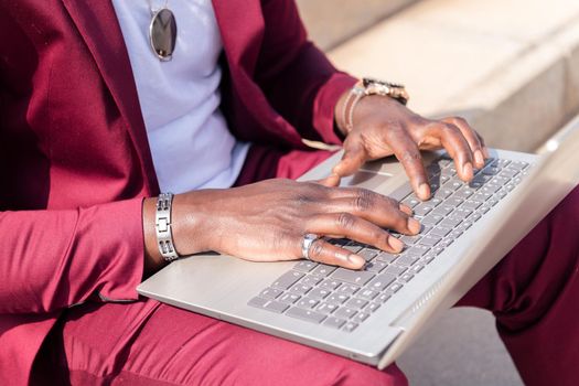 detail of the hands of an unrecognizable african man working with his laptop computer sitting on a staircase in the city, concept of technology and remote work