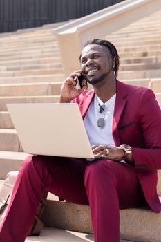 vertical photo of a smiling african businessman working with computer laptop and phone sitting on city stairs, technology and remote work concept