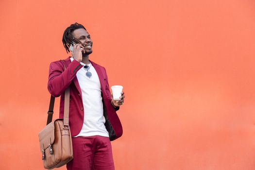 stylish african businessman dressed in suit with briefcase and coffee talks on smart phone in front of red background, copy space for text, concept of technology and communication