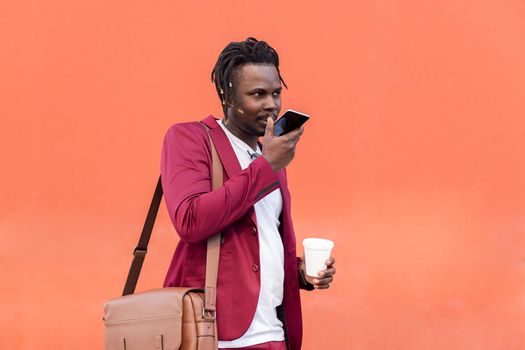 stylish black businessman dressed in suit with briefcase and coffee records a voice message on his smart phone in front of red background, copy space for text, concept of technology and communication
