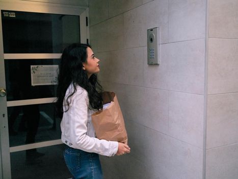 Side view of a young woman holding an eco-friendly bag stands in front of a building intercom doorbell visiting her friend.