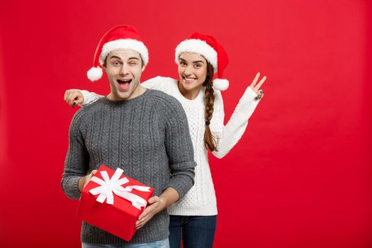 Christmas Concept - Young woman covering man's eyes with hand and giving surprise big gift. Isolated on Red background.