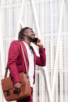 vertical photo of a smiling black businessman with briefcase walks through the city financial downtown talking on phone, concept of technology and communication, copy space for text