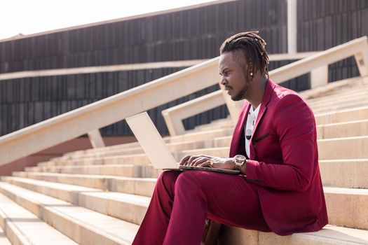stylish black businessman works with his laptop sitting on a staircase in the city, technology and remote work concept
