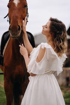 Beautiful girl in a white sundress next to a horse on an old ranch.