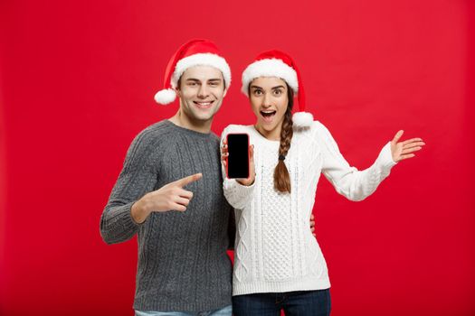 Christmas concept - Happy young couple in christmas sweaters pointing finger with mobile phone.