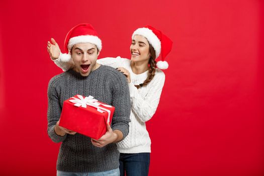 Christmas Concept - Young woman covering man's eyes with hand and giving surprise big gift. Isolated on Red background.