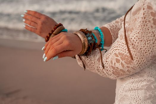 Model in boho style in a white long dress and silver jewelry on the beach. Her hair is braided, and there are many bracelets on her arms